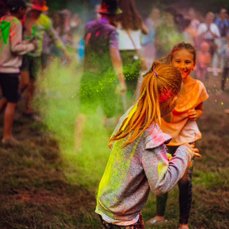 Festiwal kolorów, dziewczynki obrzucają się kolorowym proszkiem, w tle tłum ludzi w kłębach dymu.