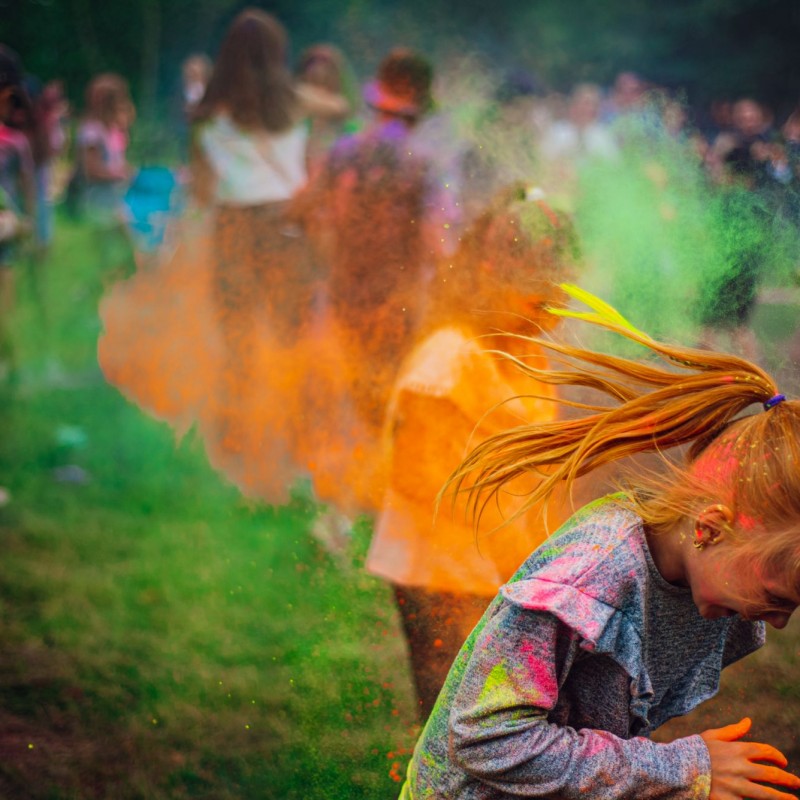 Festiwal kolorów, dziewczynka próbuje uniknąć uderzenia pomarańczowego proszku, w tle grupa ludzi w kłębach wielobarwnego dymu.