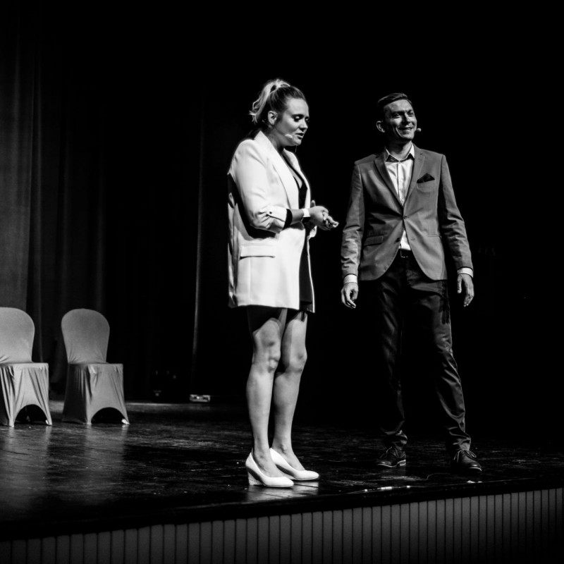 Czarno-białe zdjęcie na nim mężczyzna i kobieta ubrani na galowo stoją na scenie widowiskowej.