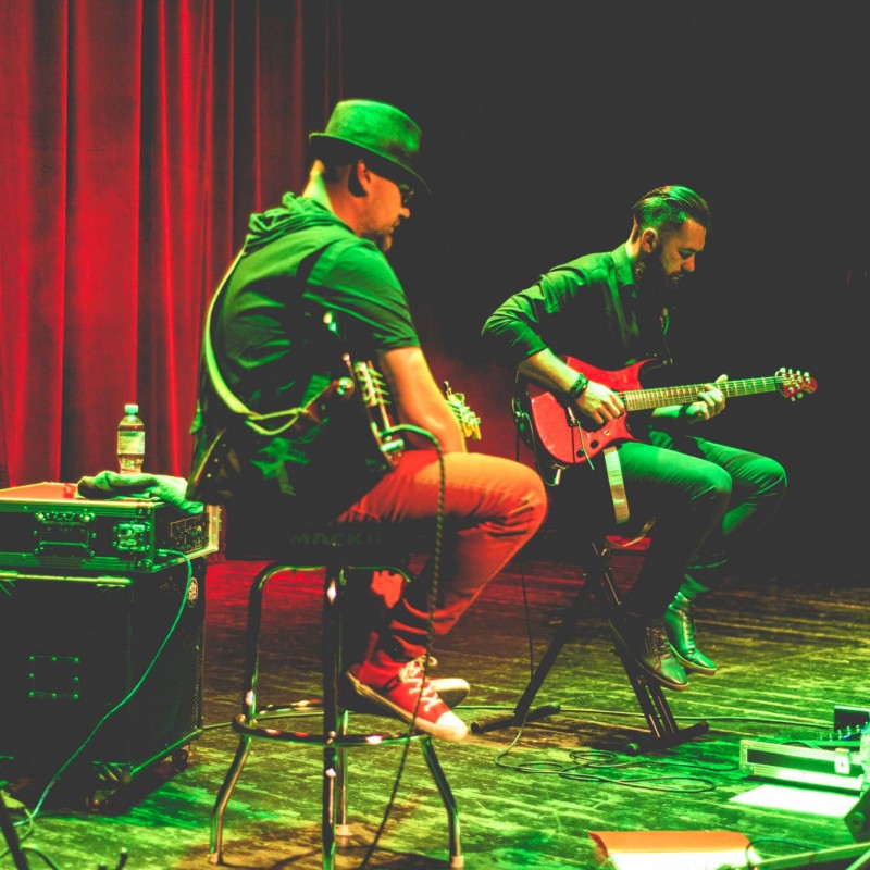 Dwóch gitarzystów siedzi na stołkach na scenie oświetlonej na zielono i czerwono podczas występu.