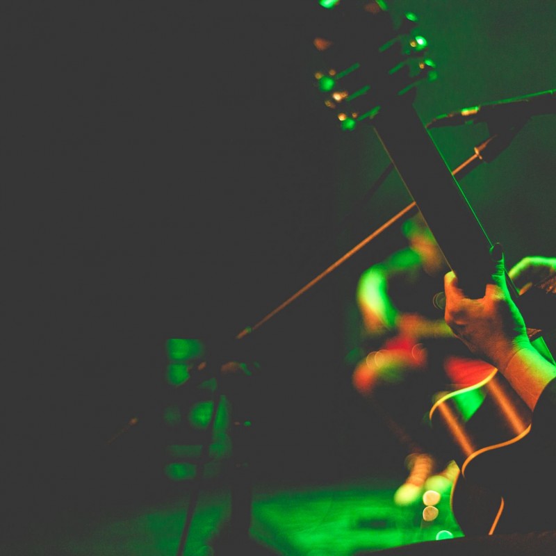 Zbliżenie na rękę gitarzystki trzymającej gryf, scena oświetlona jest w kolorach zieleni i czerwieni.