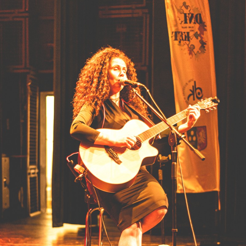 Kobieta z gitarą i długimi, kręconymi włosami siedzi na krześle gra i śpiewa do mikrofonu.