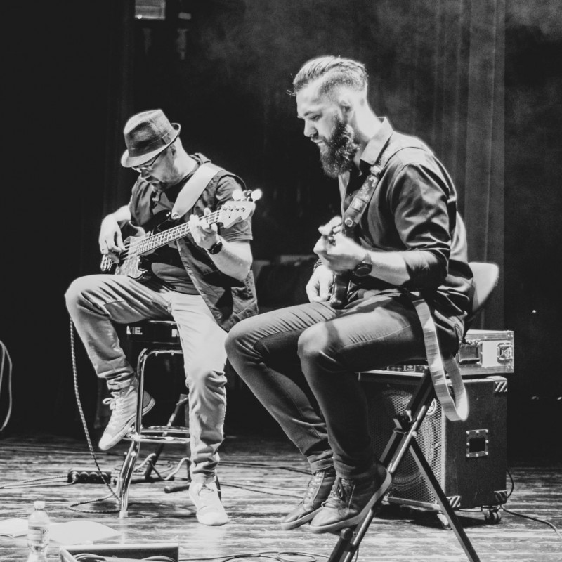 Czarno-białe zdjęcie, dwóch gitarzystów gra na gitarze siedząc na krzesłach na sali widowiskowej.