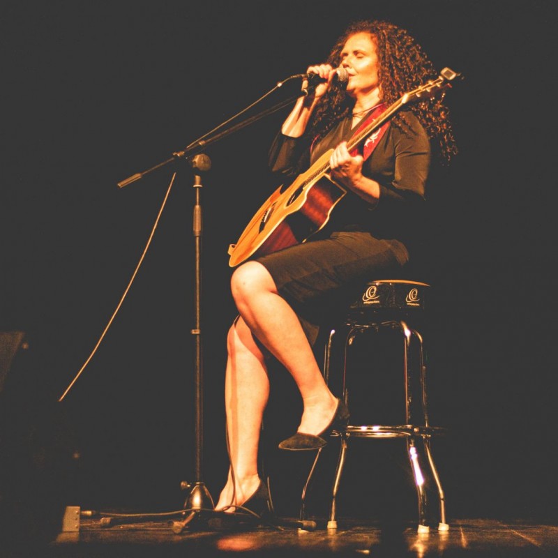 Kobieta z gitarą siedzi na krześle śpiewając do mikrofonu, obok niej gitarzysta gra na instrumencie.