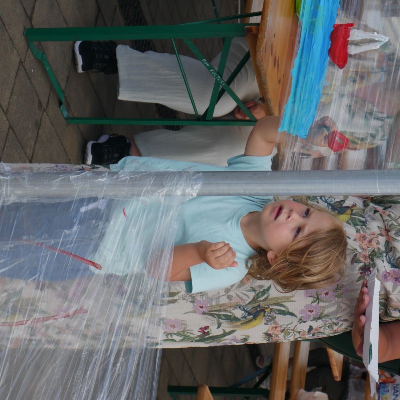 Dziecko w skupieniu maluje na rozpoztartej między dwoma słupkami przeźroczystej folii.