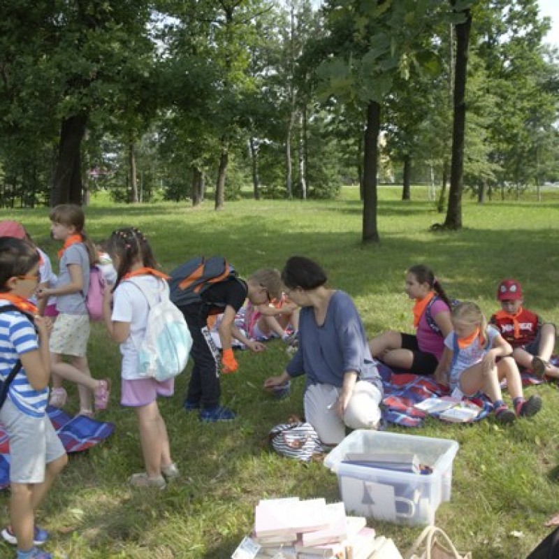 Dzieci w obecności pani instruktorki siedzą na kocach w parku.