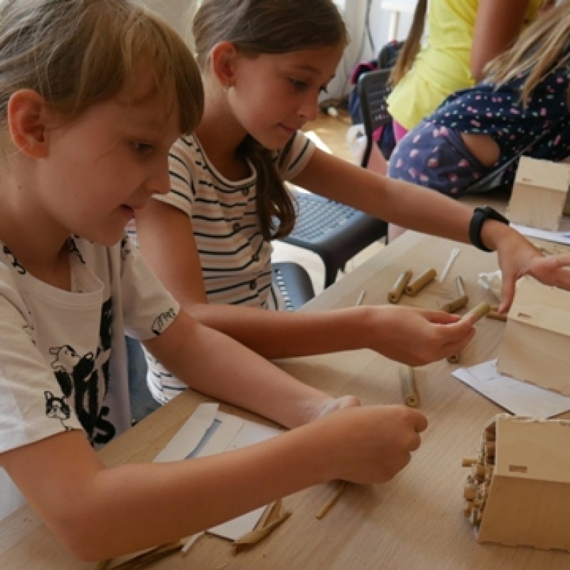 Dzieci na zajęciach plastycznych tworzą za pomocą kleju domek z drewna.