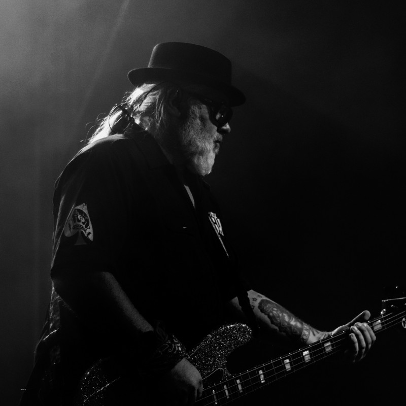 Czarno-białe zdjęcie na nim starszy mężczyzna z brodą i czapką grający na gitarze w kłębach dymu.