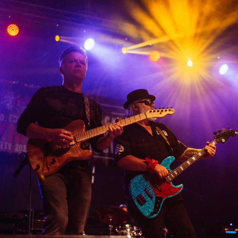 Dwóch gitarzystów podczas koncertu w kolorowym świetle reflektorów.