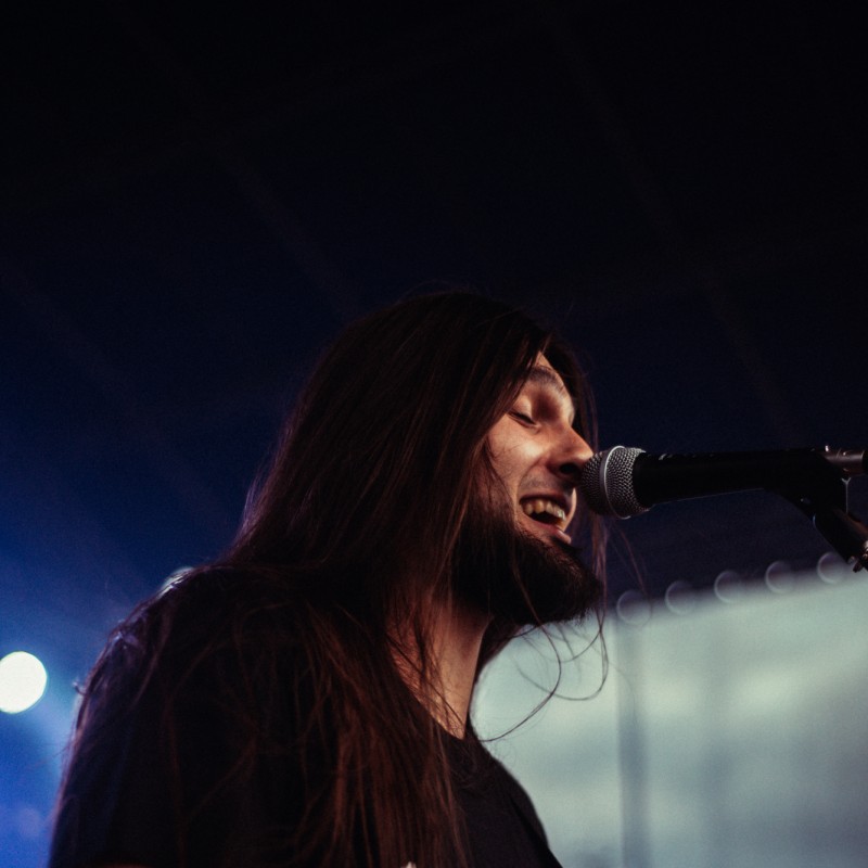 Mężczyzna z brodą i długimi włosami śpiewa do mikrofonu podczas występu.