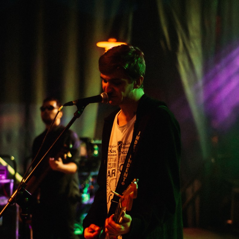 Mężczyzna w kolorowych światłach na scenie gra na gitarze i śpiewa do mikrofonu.