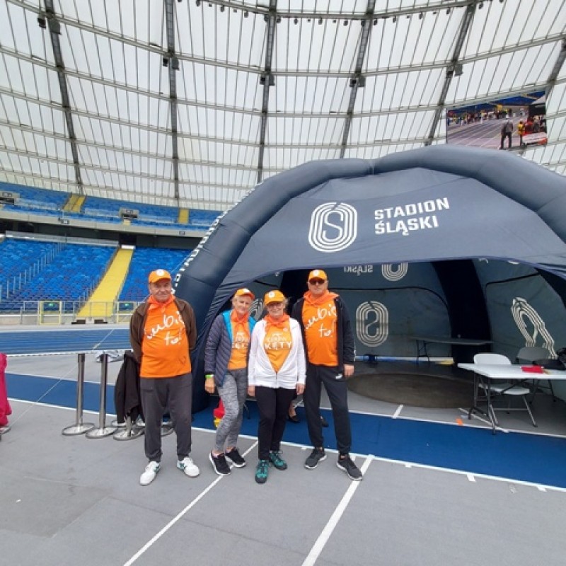 Czwórka ludzi w pomarańczowych koszulkach stoi przed szarym namiotem festiwalowym z napisem „Stadion Śląski”.
