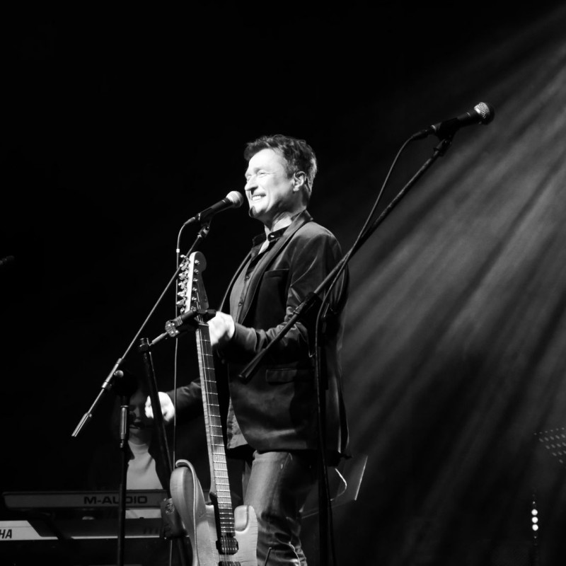 Na środku sceny, przed mikrofonem, stoi uśmiechnięty mężczyzna, a obok niego znajduje się gitara. Zdjęcie jest czarno-białe.