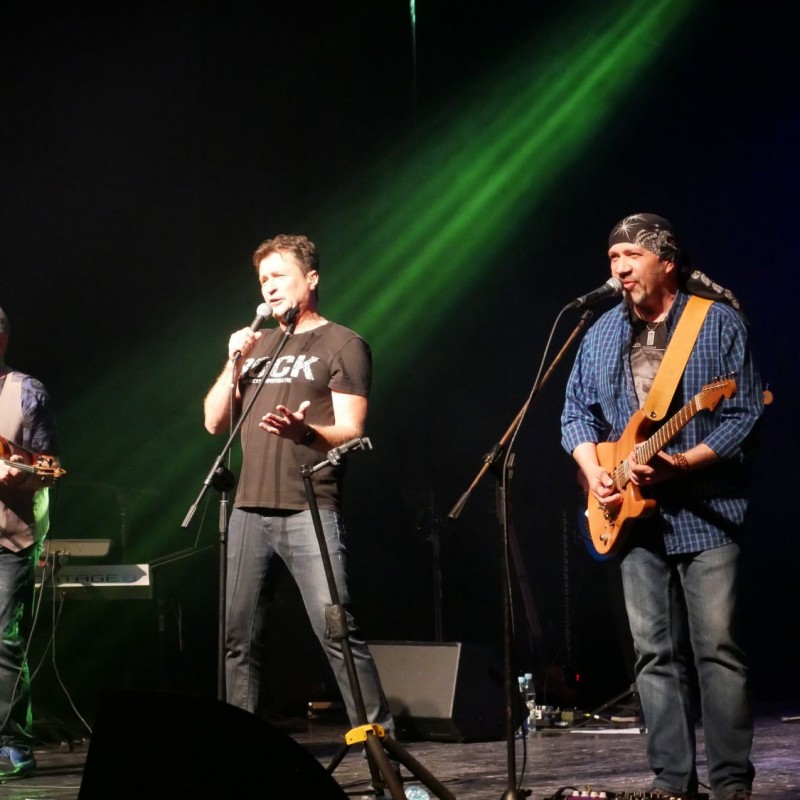 Trzech panów stoi przed mikrofonami. Mężczyzna po lewej trzyma w dłoniach skrzypce, natomiast ten po prawo – gitarę.