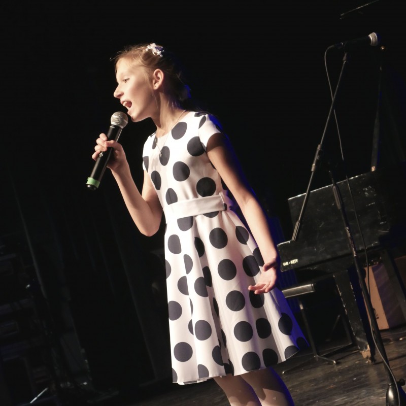 Dziewczynka w sukience w kropki śpiewa do mikrofonu na scenie widowiskowej.