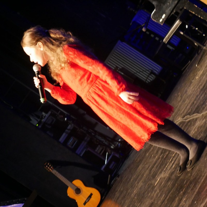 Kobieta ubrana w czerwoną sukienkę śpiewa do mikrofonu na scenie widowiskowej.