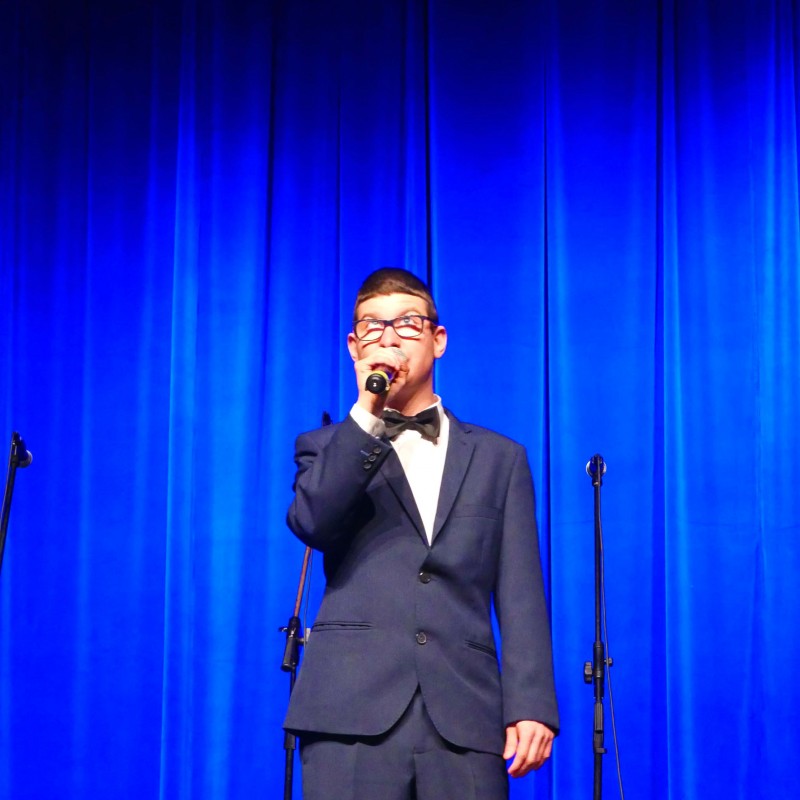 Mężczyzna w garniturze śpiewa do mikrofonu na scenei widowiskowej.