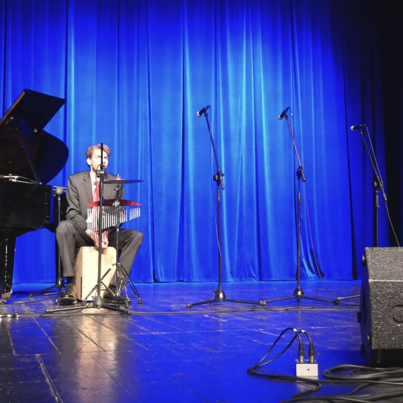 Mężczyzna w garniturze śpiewa do mikrofonu przy akompaniamencie kahonu oraz pianina.