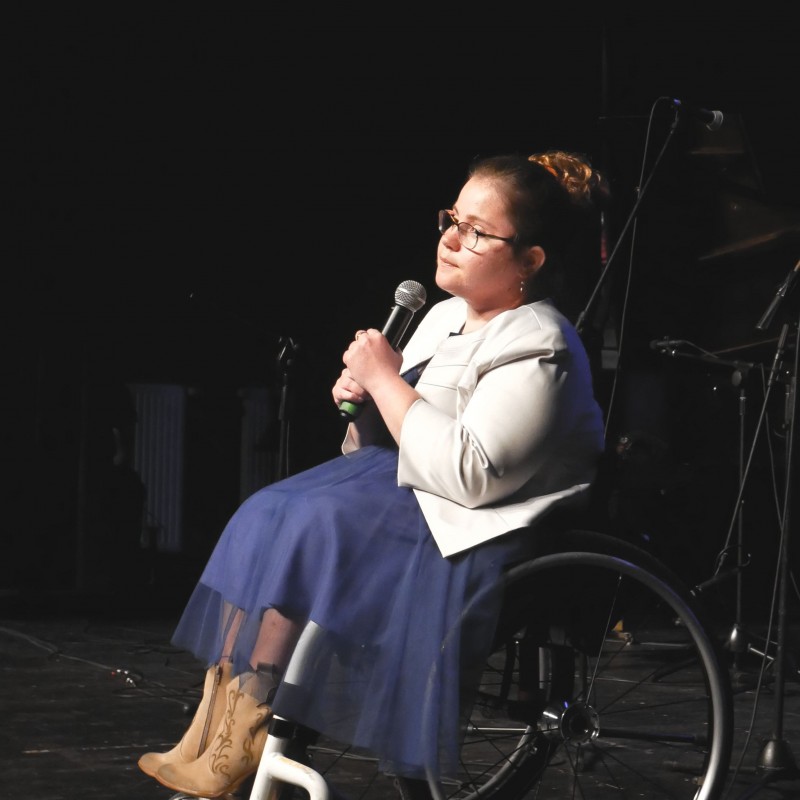 Kobieta na wózku podczas występu na scenie, w dłoni trzyma mikrofon.