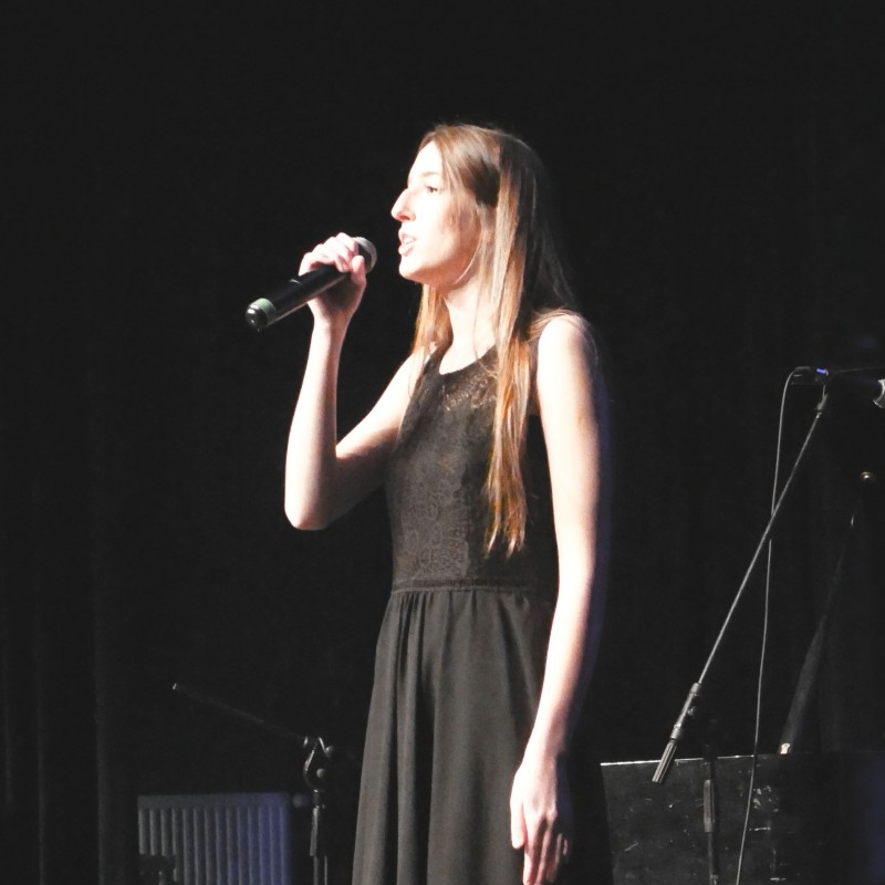Kobieta ubrana w czarną sukienkę śpiewa do mikrofonu na scenie w świetle reflektorów.