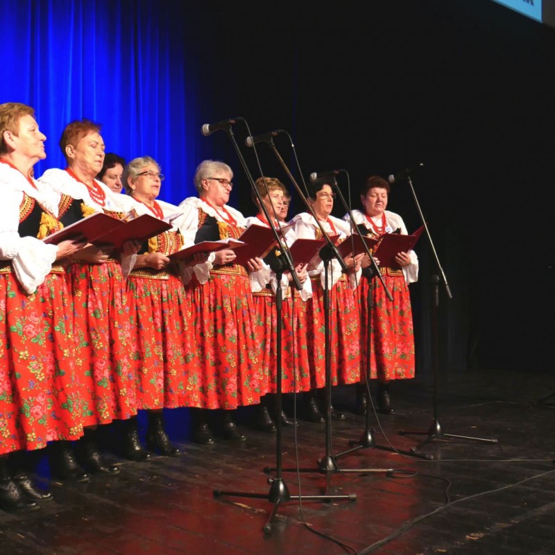 Chór kobiet podczas występu na scenie ubrany w stroje regionalne z czerwonymi, kwiecistymi spódnicami przy akompaniamencie akordeonu.