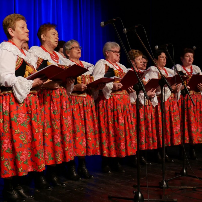 Grupa kobiet ustawionych na scenie ubranych w stroje regionalne w czerwonych kwiecistych spódnicach trzyma śpiewniki w rękach.