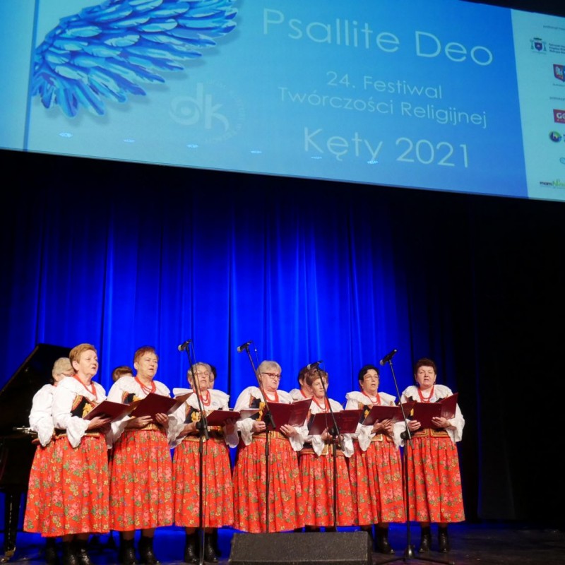 Chór kobiet podczas występu na scenie ubrany w stroje regionalne z czerwonymi, kwiecistymi spódnicami przy akompaniamencie akordeonu.