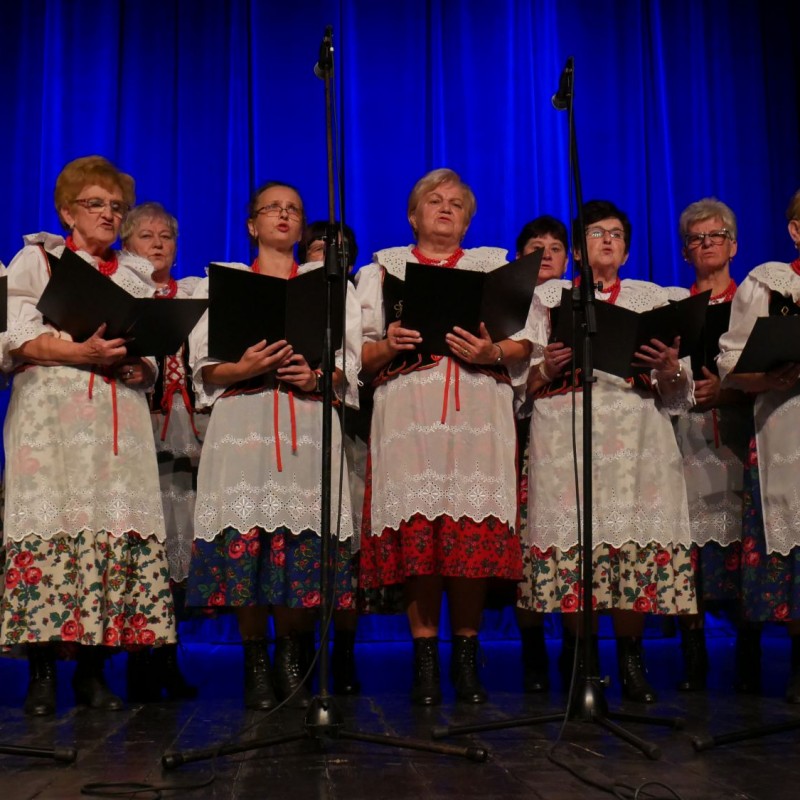 Kobiecy chór podczas występu na scenie trzymający w dłoniach pulpity w kolorowych strojach regionalnych.