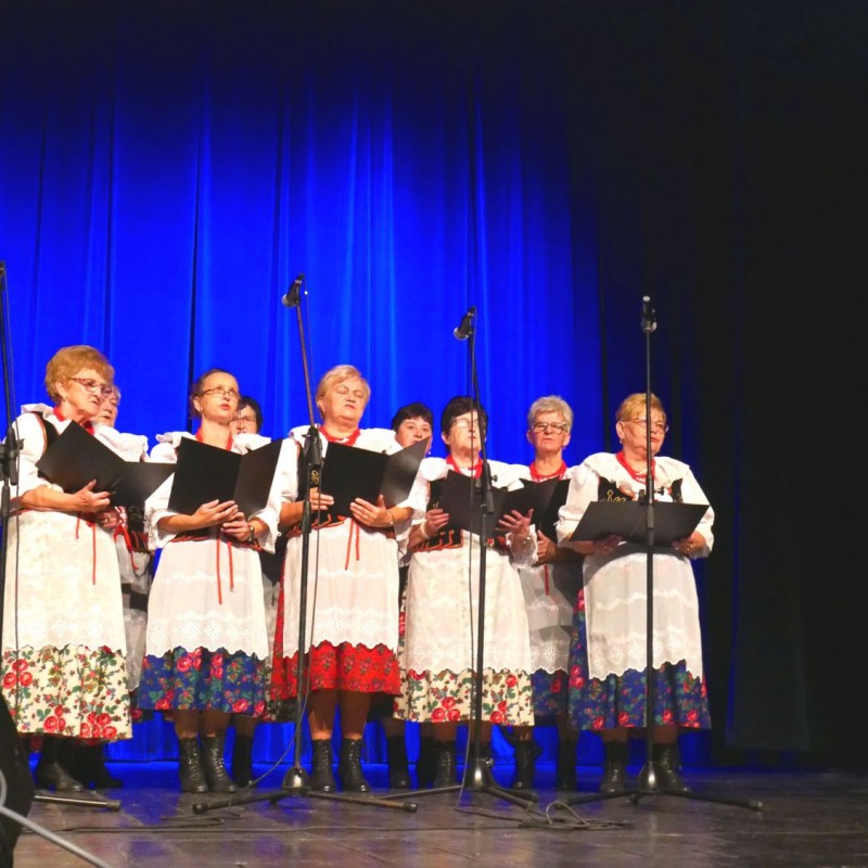 Chór kobiet śpiewa na scenie z pulpitami w rękach rzy akompaniamencie akordeonu, ubrane są w kolorowe stroje regionalne.