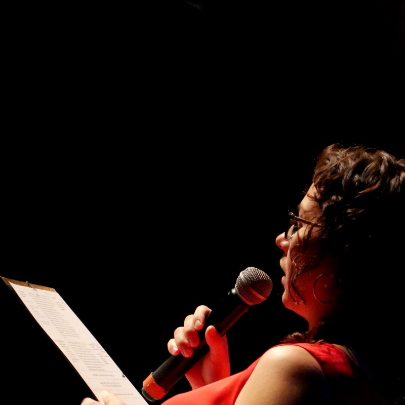 Konferansjerka w czerwonej sukience czyta z pulpitu, który trzyma w dłoni do mikrofonu. 