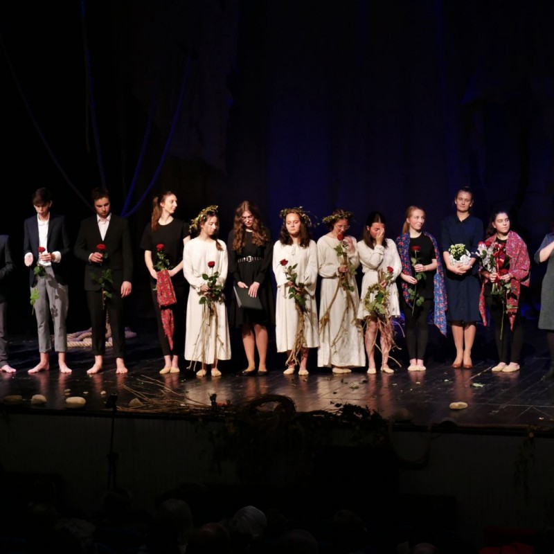 Na scenie grupa ludzi, w różnych strojach. Każdy z nich trzyma w rękach różę. Po prawej stronie kobieta w eleganckiej sukience, która mówi do trzymanego w dłoni mikrofonu. 