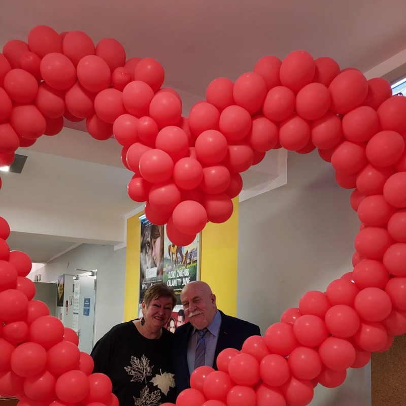 Dwójka seniorów pozuje za ogromnym sercem stworzonym z czerwonych balonów.