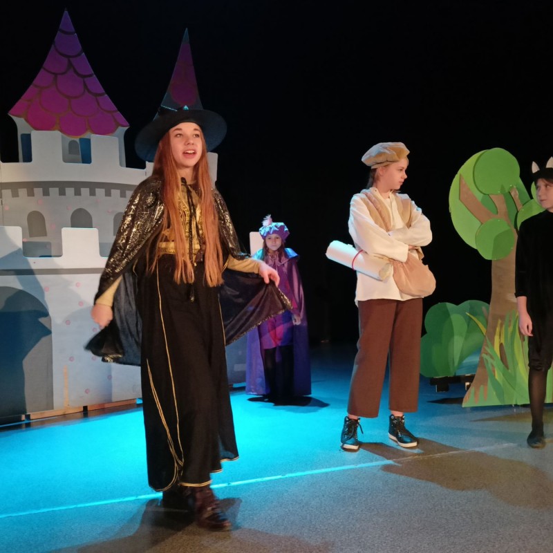 Dziewczyna przebrana za czarnoksiężnika w trakcie recytacji stoi na scenie, za nią scenografia zamku i lasu, obok inni wykonawcy ubrani w kostiumy.