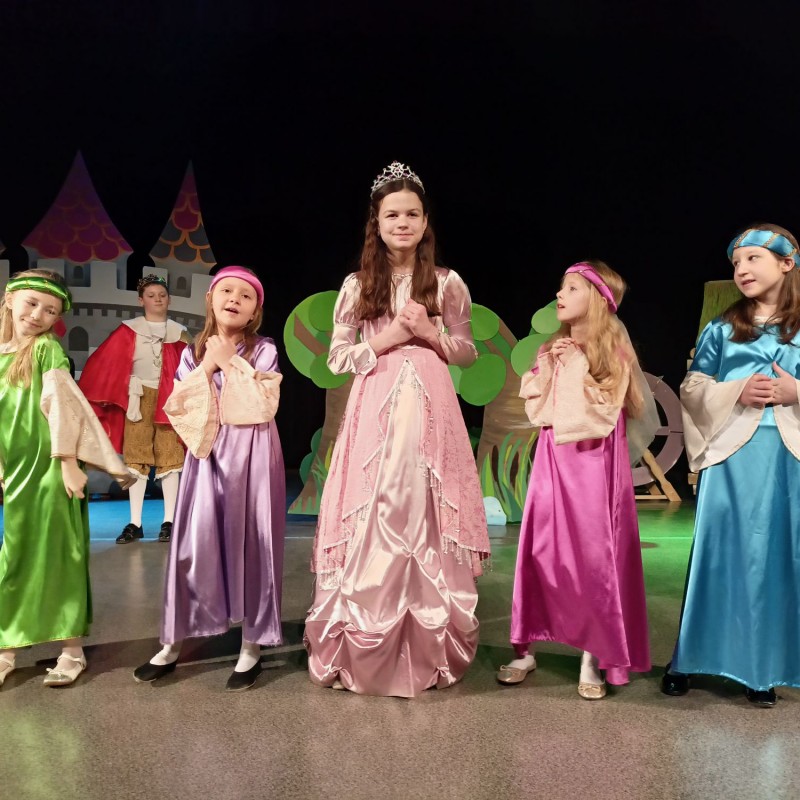 Dziewczyna przebrana za księżniczkę stoi na scenie w obecności czterech dam dworu, w tle król.