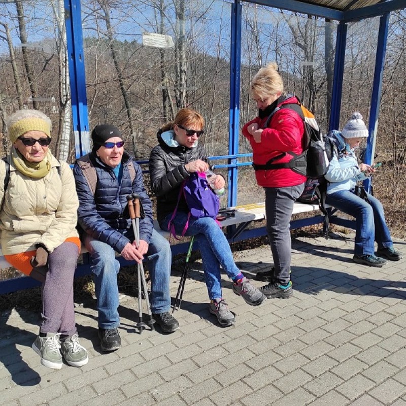 Seniorzy w górskich strojach z kijkami siedzący na przystanku czekający na start wycieczki.