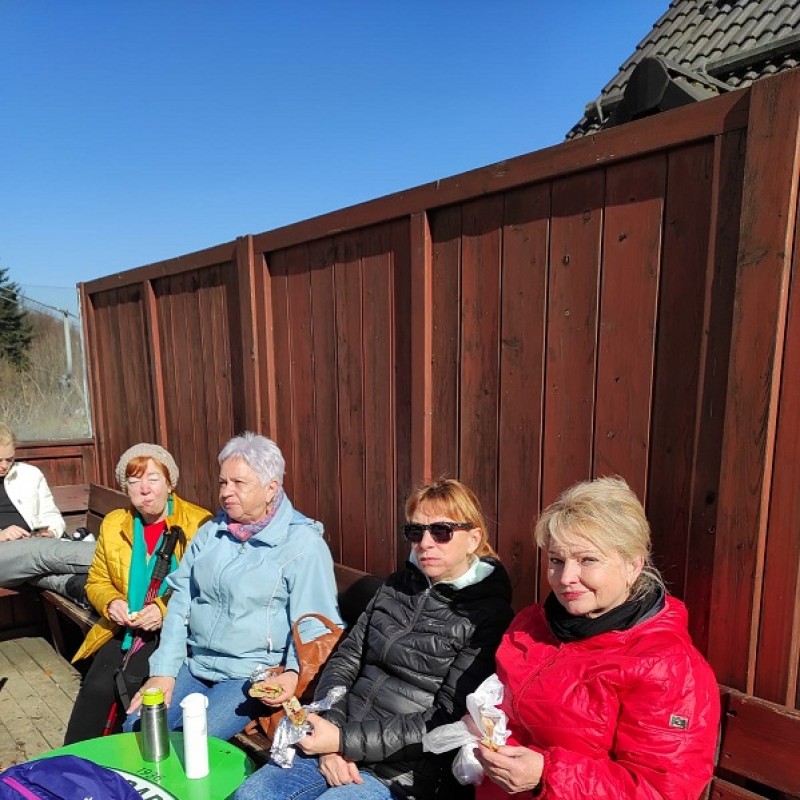 Uczestnicy wycieczki siedzący w słońcu na ławce i jedzący.