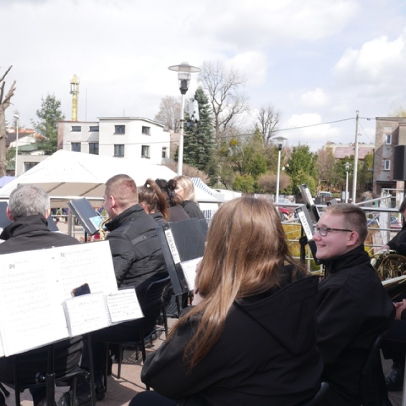 Orkiestra siedząca przed domem kultury podczas jarmarku, przed nimi pulpity z nutami.