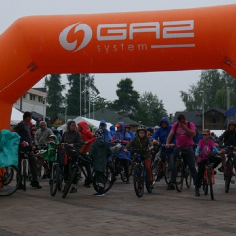 Start rajdu rowerowego przed domem kultury, uczestnicy w kapokach przeciwdeszczowych stoją na rowerach pod dmuchanego pomarańczowego balonu.