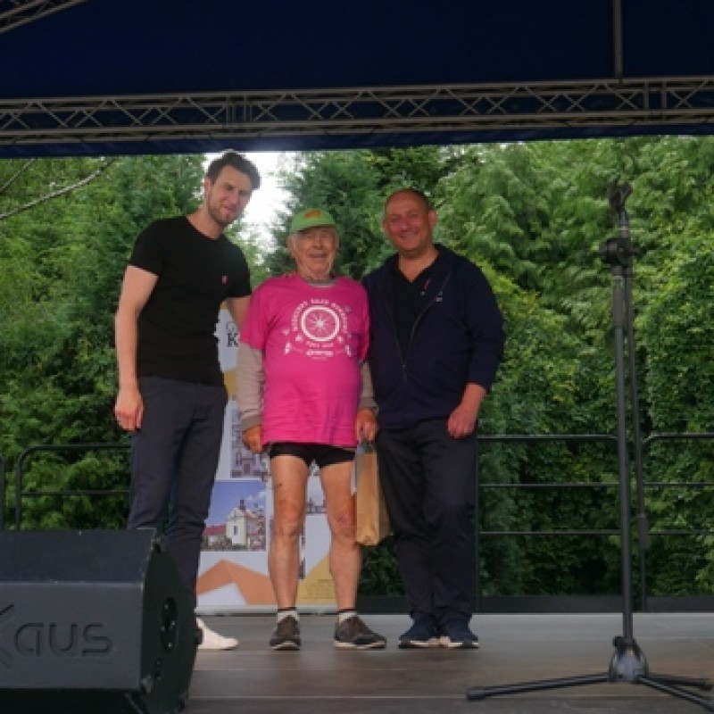 Scena plenerowa, burmistrz gminy Kęty i wicedyrektor domu kultury pozują ze starszym panem otrzymującym nagrodę w różowej rajdowej koszulce.