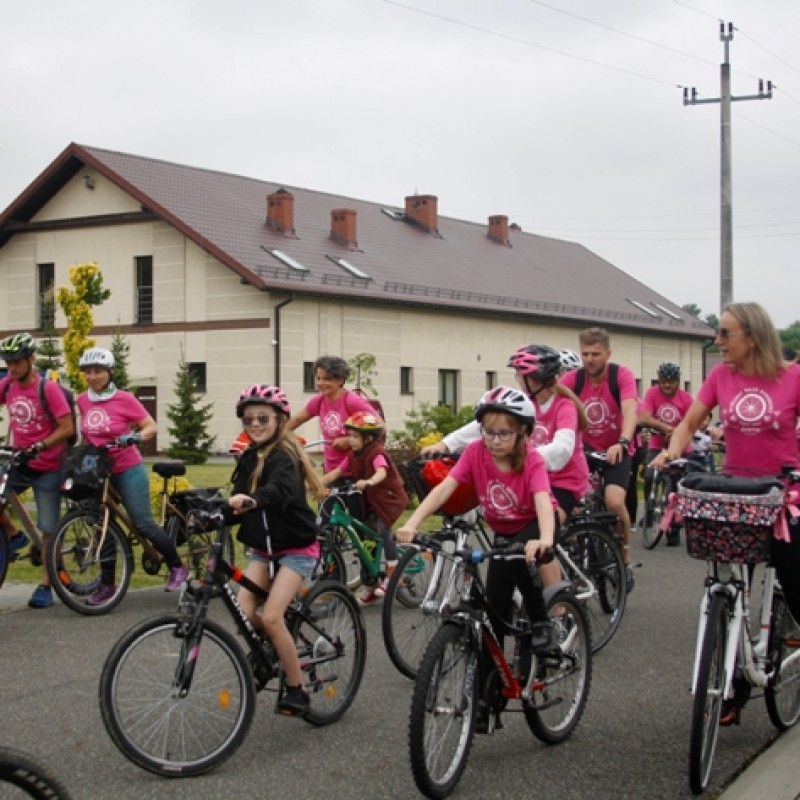 Rajd rowerowy ludzi ubranych w różowe koszulki w trakcie jazdy na ulicy, w tle duży budynek.