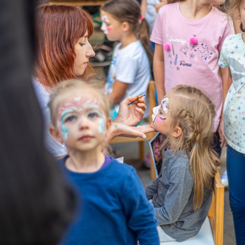 Fot. Łukasz Kuc/Dziewczynka ma wykonywany malunek na twarzy przez kobietę, stoją na około niej inne dzieci. 