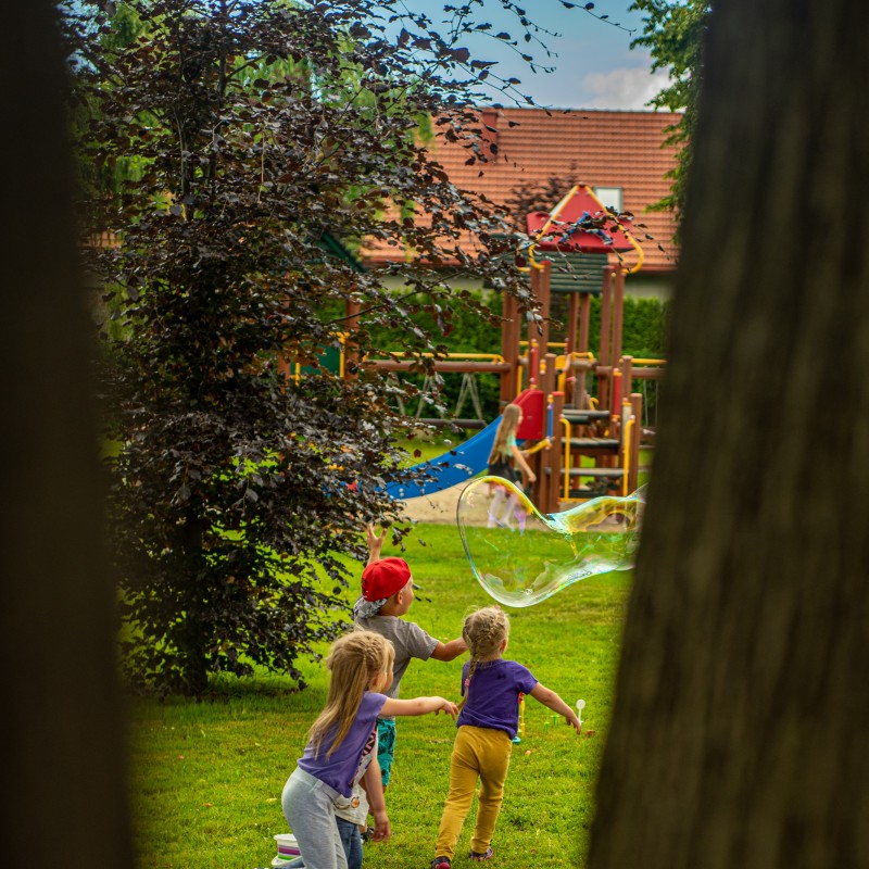 Fot. Łukasz Kuc/Zza drzewa widać dzieci biegnące za ogromną mydlaną bańką obok chusty Klanzy, w tle plac zabaw.