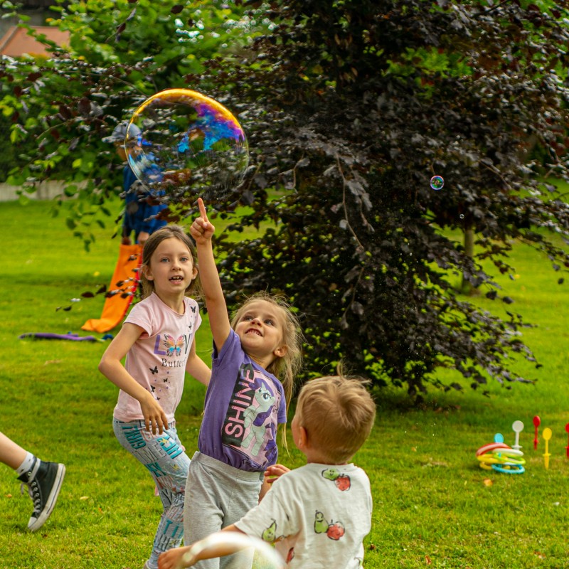 Fot. Łukasz Kuc/Trójka dzieci biega za bańkami mydlanymi, jedno z nich prawie dotyka palcem jedną z baniek