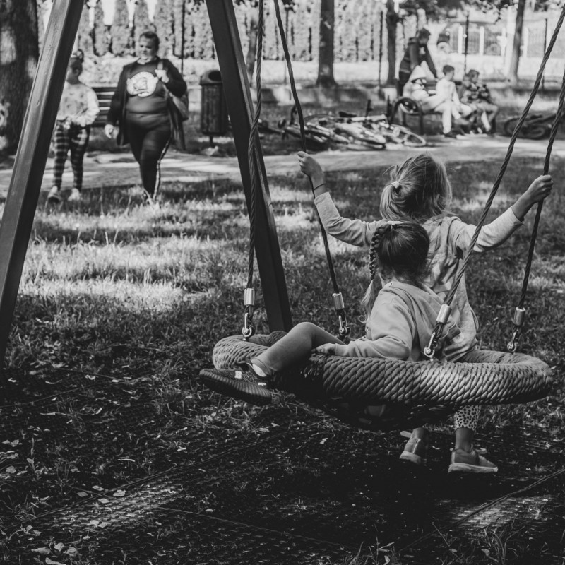 Fot. Łukasz Kuc/Czarno białe zdjęcie, w parku na huśtawce dwie dziewczynki, z tyłu mam z dzieckiem idą w ich stronę.