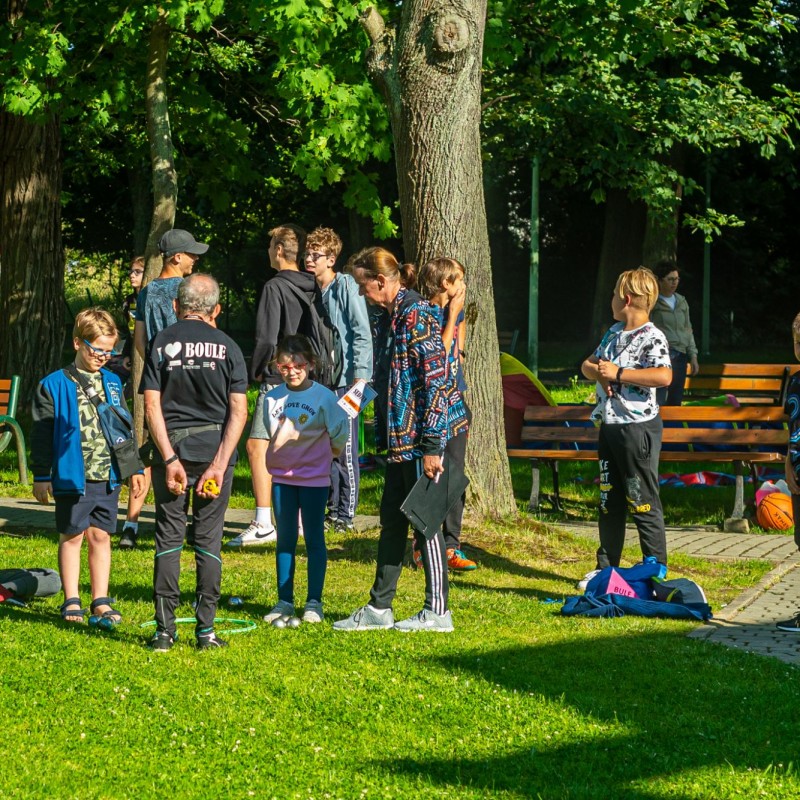 Fot. Łukasz Kuc/Grupa dzieci i dorosłych stoi w słoneczny dzień w parku. Starszy pan tłumaczy im zasady gry z boule.