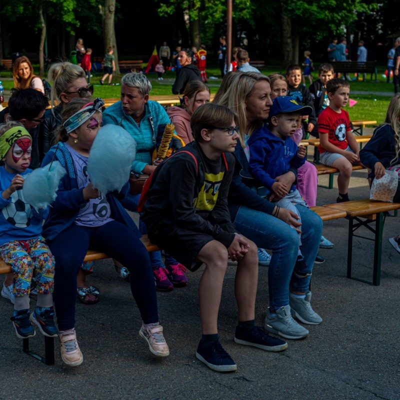 Fot. Łukasz Kuc/Na placu w parku na ławkach ustawionych rzędowo siedzą dorośli i dzieci i przyglądają się czemuś.