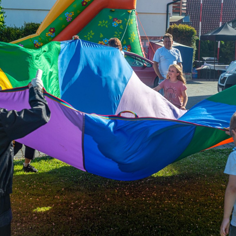 Fot. Łukasz Kuc/Dzieci bawią się kolorową chustą Klanzy na świeżym powietrzu, w tle dmuchana zjeżdżalnia i domy.
