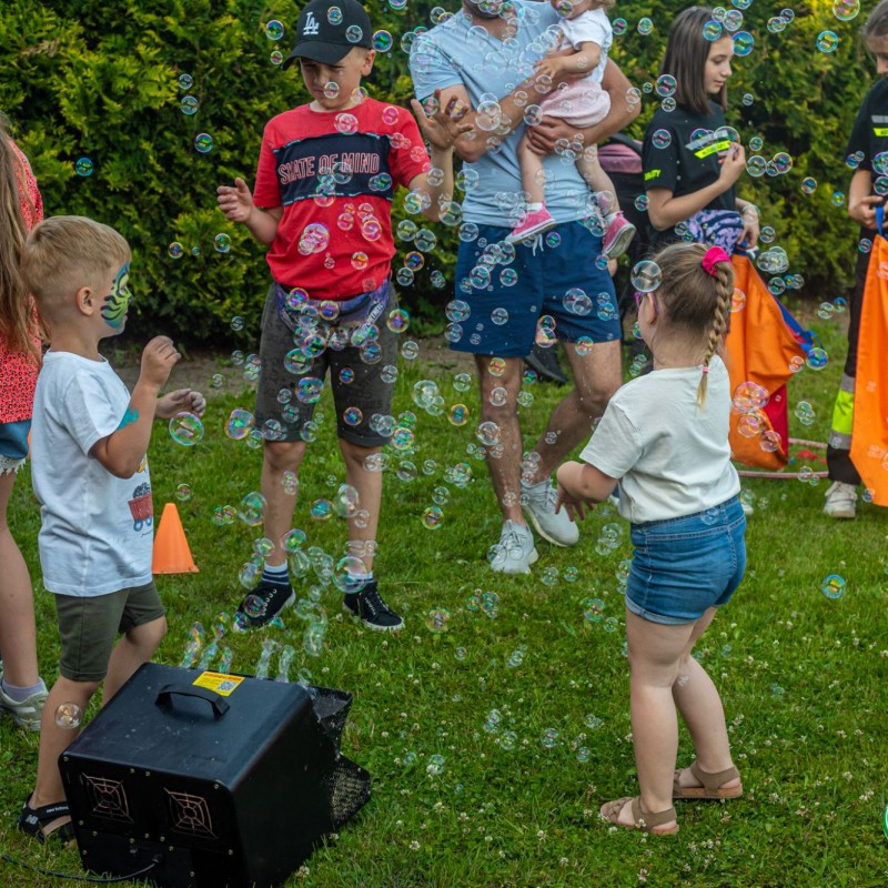 Fot. Łukasz Kuc/Dzieci bawią się w otoczeniu baniek mydlanych produkowanych przez maszynkę. Znajdują się na trawie w otoczeniu krzewów.