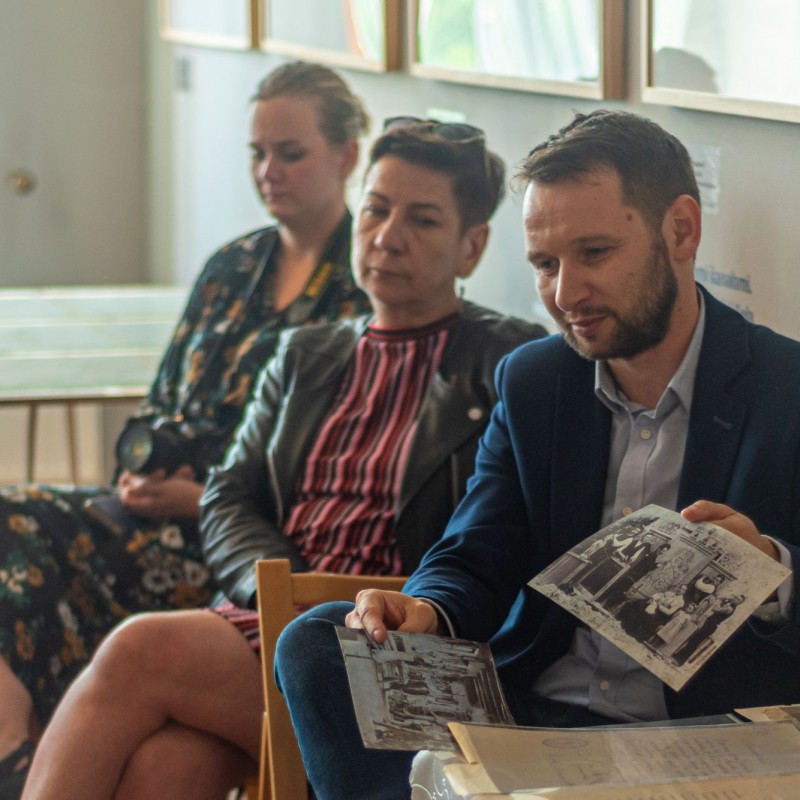 Fot. Łukasz Kuc/Mężczyzna siedzi przy biurku trzymając w dłoniach czarno-białe fotografie, obok niego siedzą dwie kobiety.