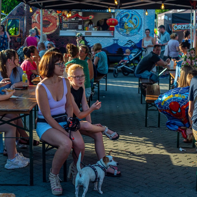 Fot. Łukasz Kuc/Ludzie siedzący przy stolikach na placu domu kultury, w tle food trucki, a z przodu kobieta z małym psem.